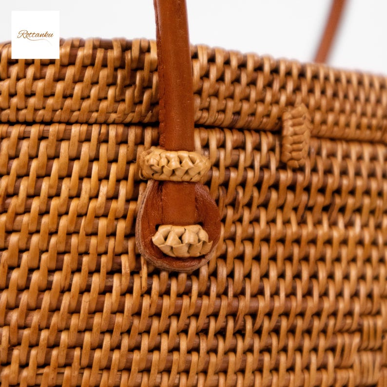 May include: Close-up of a woven rattan handbag with a brown leather handle. The bag is made of tightly woven, light brown rattan. The handle is attached with a decorative woven detail. The bag has a natural, earthy aesthetic.