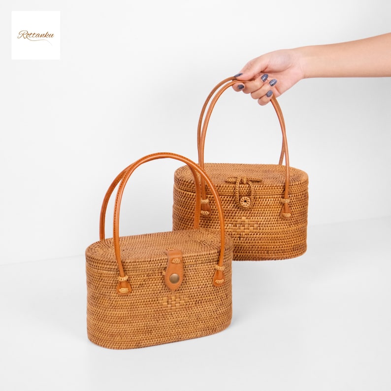 May include: Two oval-shaped rattan handbags with leather handles. One is being held. The bags are light brown and have a woven texture. Each bag has a clasp closure. The bags are a natural, rustic style.