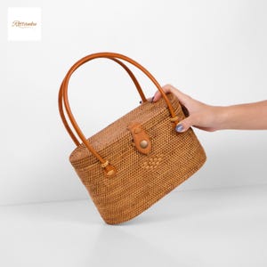 May include: A rectangular woven rattan handbag with a brown leather handle and a leather clasp. The bag is a light brown colour and has a natural, textured appearance. The bag is held by a person's hand.