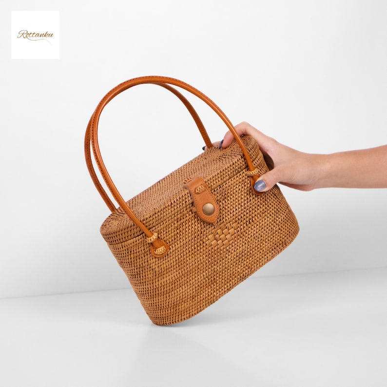 May include: A rectangular woven rattan handbag with a brown leather handle and a leather clasp. The bag is a light brown colour and has a natural, textured appearance. The bag is held by a person's hand.