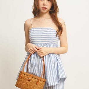 May include: A blue and white striped romper with thin straps. The outfit is paired with a rectangular woven rattan handbag with brown leather handles and accents. The overall style is casual and summery.