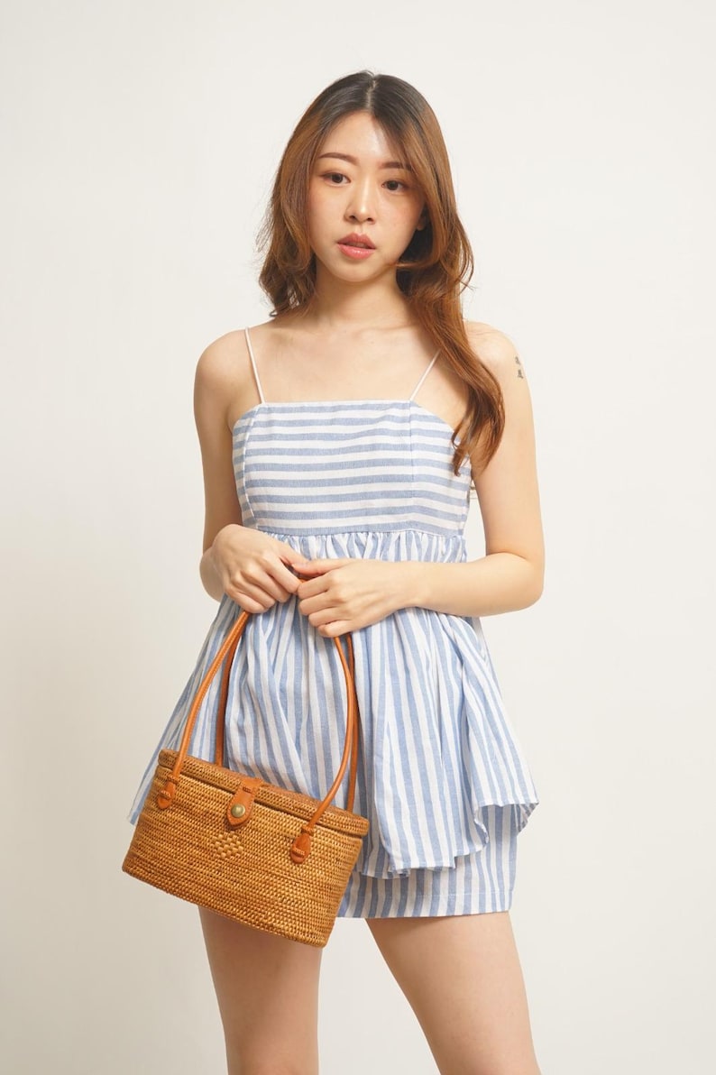 May include: A blue and white striped romper with thin straps. The outfit is paired with a rectangular woven rattan handbag with brown leather handles and accents. The overall style is casual and summery.