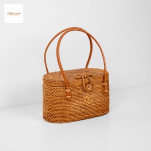 May include: Oval-shaped woven rattan handbag with a brown leather handle and clasp. The bag features a rectangular shape with rounded edges and a woven pattern. The handle is arched, and the clasp is centered on the front. The bag has the text "Rottanku" in the upper left corner.