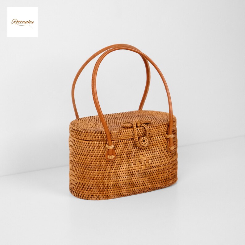 May include: Oval-shaped woven rattan handbag with a brown leather handle and clasp. The bag features a rectangular shape with rounded edges and a woven pattern. The handle is arched, and the clasp is centered on the front. The bag has the text "Rottanku" in the upper left corner.