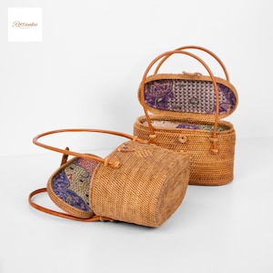 May include: Two woven rattan handbags with leather handles. One bag is open, revealing a patterned fabric lining in shades of purple, red, and green. The other bag is closed. The bags are light brown in colour.