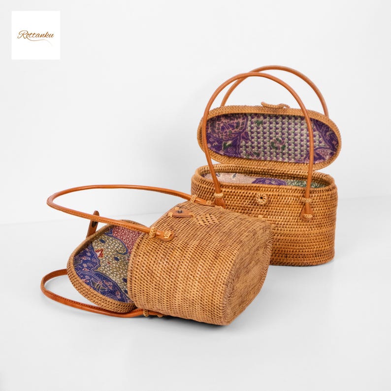 May include: Two woven rattan handbags with leather handles. One bag is open, revealing a patterned fabric lining in shades of purple, red, and green. The other bag is closed. The bags are light brown in colour.