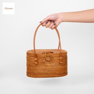 May include: A woven rattan handbag with a brown leather handle and a clasp closure. The oval-shaped bag is held by a hand against a white background. The bag measures approximately 25 cm in length.