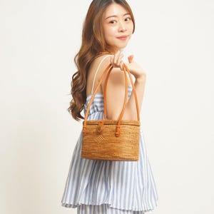 May include: A rectangular, woven rattan handbag with brown leather handles and accents. The bag has a button closure and is held by a person wearing a blue and white striped dress. The bag is a natural tan colour.