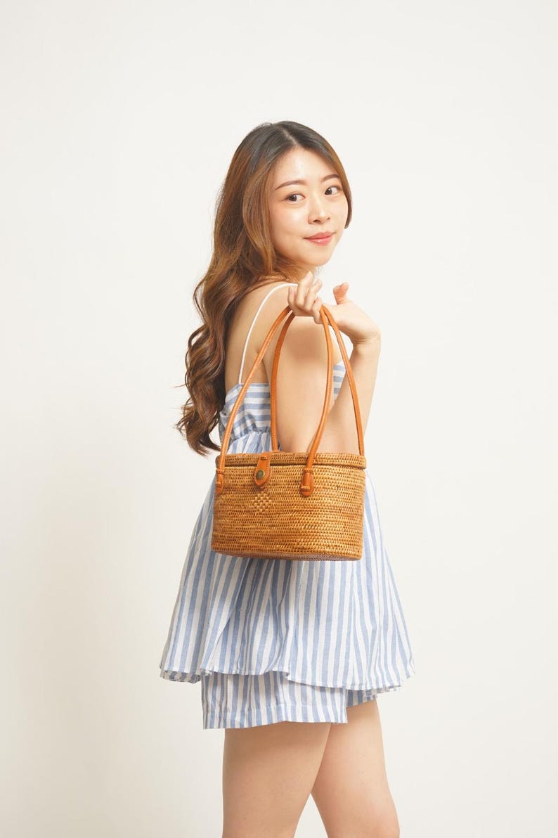 May include: A rectangular, woven rattan handbag with brown leather handles and accents. The bag has a button closure and is held by a person wearing a blue and white striped dress. The bag is a natural tan colour.