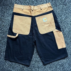 May include: Black and tan work shorts with multiple pockets. The shorts have a tan-colored waistband and back pockets, with a small logo patch. The front and the majority of the back are black. The shorts are made of a durable material.