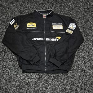 Può includere: Una giacca nera con zip caratterizzata dal logo McLaren in bianco e giallo. La giacca include varie toppe ricamate, tra cui un emblema McLaren F1 con il testo "There is no finish line" e una toppa "1963" con un grafico di un casco da corsa.