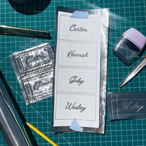 May include: A craft project in progress, featuring a sheet of foil with embossed names: "Carter", "Hannah", "Gaby", and "Wesley". Tools include a pencil, scissors, tape, and a burnishing tool, all on a green grid cutting mat.