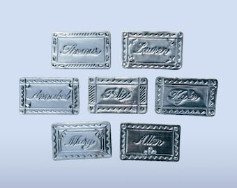 Peut inclure: Sept plaques rectangulaires en métal argenté, chacune dotée d'une bordure décorative et d'un nom gravé. Les noms affichés sur les plaques individuelles sont Thomas, Lauren, Annabel, Felix, Kyler, Mary et Allen, tous écrits en écriture cursive.