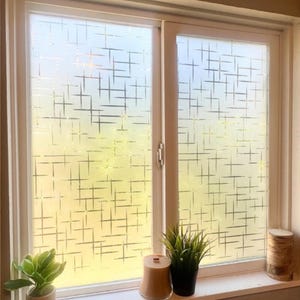 May include: A window with a white frame and a frosted glass design featuring a pattern of intersecting lines. The window is partially open, and the interior is visible. A small plant, a candle, and a potted plant sit on the windowsill.
