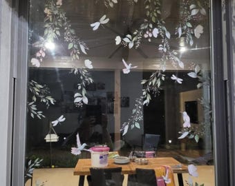May include: A glass window adorned with floral and fauna decals. The design includes white flowers, green leaves, dragonflies, and birds. A wooden table and chairs are visible through the window, creating a nature-inspired aesthetic.