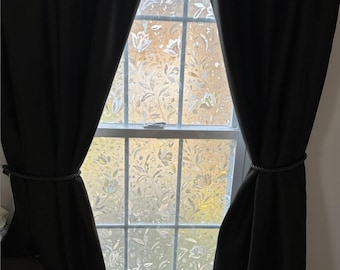 May include: A window with a floral design, framed by dark curtains tied back with rope. The window's glass has a frosted appearance with a pattern of flowers and leaves. The curtains are a solid, dark color.