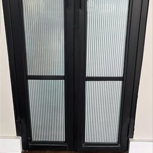 May include: Black double doors with vertical, frosted glass panels. The doors have a simple, modern design with black frames and a textured, ribbed glass pattern. The doors are closed and appear to be interior doors.