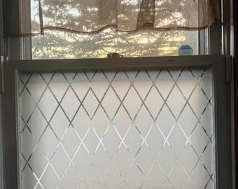 May include: A window with a diamond pattern etched into the glass. The window is framed in a neutral color and has a sheer curtain at the top. A Ring security device is visible in the upper right corner.