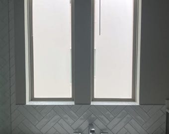 May include: A bathroom scene featuring a white bathtub, a chrome faucet, and a herringbone-patterned tile backsplash. Above the tub are two frosted windows with blinds. The walls are painted a light gray color.