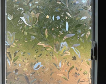 May include: A window with a decorative floral pattern. The design features stylized flowers and leaves in shades of white, blue, and green, creating a frosted glass effect. The window is framed in white and has a view of a blurred green background.