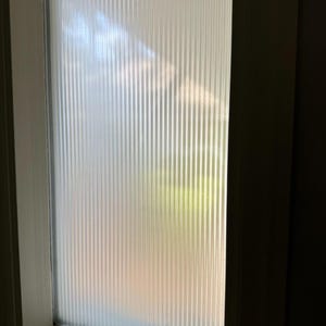 May include: A close-up of a frosted glass panel with vertical, textured lines. The panel is set within a white frame, with a glimpse of the outside visible through the glass. The light filters through, creating a soft, diffused effect.