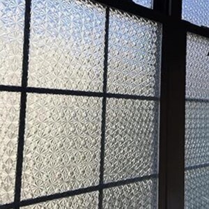 May include: Close-up of a window with textured glass panels. The glass has a repeating pattern of small, raised floral shapes. Black frames divide the window into rectangular sections, creating a grid-like structure. The light shining through the glass gives it a bright appearance.