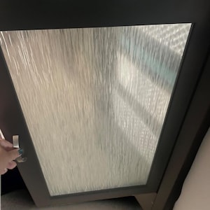 May include: A dark brown cabinet door with a textured glass panel. The door is slightly ajar, revealing a glimpse of the interior. A hand is reaching for the silver handle.