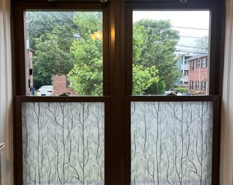 May include: A double-hung window with a dark brown frame. The window panes feature a frosted film with a pattern of tree branches and leaves. The view outside shows green trees, a brick building, and a car.