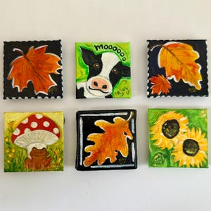 May include: Six small square paintings with autumn and farm themes. The paintings depict a cow with the word "moooooo", fall leaves, a mushroom with a frog, and sunflowers. Each painting has a black border.