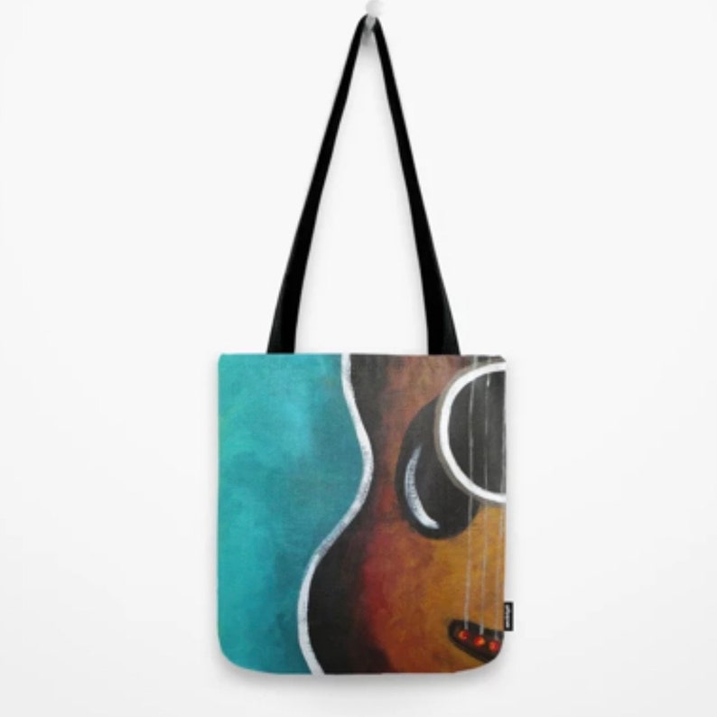 May include: Tote bag featuring a painted acoustic guitar design. The guitar is depicted in shades of brown and yellow, with black strings and a red accent. The background is a teal blue color, and the bag has black handles.
