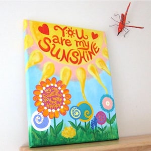 May include: A hand-painted canvas with a yellow sun and the text "You are my sunshine" in red and orange. The sun has a pink and yellow background. Below the sun are colorful flowers with white dots and the text "my only sunshine you make me happy".