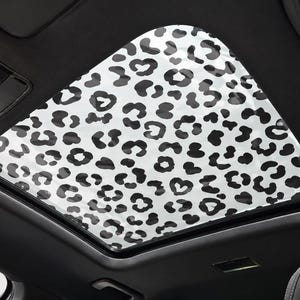 May include: A car sunroof shade featuring a white background with a repeating black leopard print pattern. The shade is installed within the rectangular frame of the vehicle's interior roof, covering the glass panel to block out sunlight.