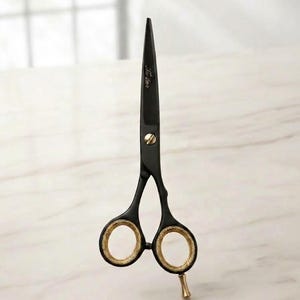 Könnte beinhalten: Schwarze Haarschneideschere mit goldfarbener Schraube, goldfarbener Fingerauflage und goldfarbenen, texturierten Innenringen. Die Klingen sind mattschwarz und weisen ein kleines eingraviertes Logo auf der oberen Klinge nahe dem Gelenk auf.