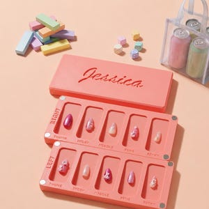 May include: A coral-colored organizer tray set for press-on nails, labeled with finger positions, featuring ten decorative nail tips. The set includes a matching coral lid with the name Jessica embossed on top, alongside pastel blocks and a clear bag.