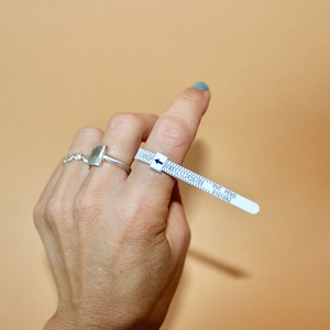 以下が含まれることがあります： 青い矢印と黒い数字が付いた白いプラスチック製のリングサイズ測定器。測定器は、銀の指輪をした指に巻かれている。測定器のテキストには「PAT. PEND. 9101693」と記載されている。