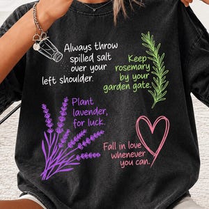 以下が含まれることがあります： 白、緑、紫、ピンクの文字とイラストが描かれた黒いTシャツ。塩入れ、ローズマリーの小枝、ラベンダーの植物、ハートのグラフィックがプリントされており、塩、ローズマリー、ラベンダー、愛に関するフレーズが添えられています。
