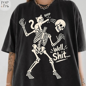 Könnte beinhalten: Ein schwarzes T-Shirt mit einer Grafik eines Skeletts mit abgetrenntem Schädel, während eine kleine Katze auf seinen Schultern sitzt und mit dem Text BOOP auf den Schädel tippt. Der Text Well, Shit... steht in weiß unter dem Skelett.