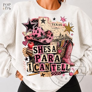 Può includere: Una felpa girocollo grigio chiaro con una stampa grafica raffigurante un cappello da cowboy rosa con macchie di mucca, uno stivale da cowboy rosa, una mela rossa, libri, matite e una mappa del Texas con il testo "SHE'S A PARA I CAN TELL" in nero e rosa.