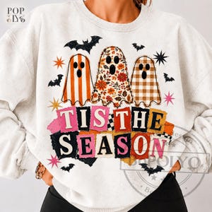 Puede incluir: Una sudadera gris claro de cuello redondo con un gráfico de tres fantasmas con diferentes patrones: rayas naranjas, estampado floral y cuadros vichy marrones. Debajo de los fantasmas, el texto TIS THE SEASON aparece en letras de colores.