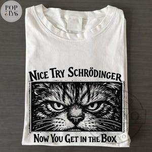 May include: White t-shirt featuring a black and white illustration of a cat's face with an angry expression. The text "Nice Try Schrödinger, Now You Get in the Box" is printed above and below the cat image.