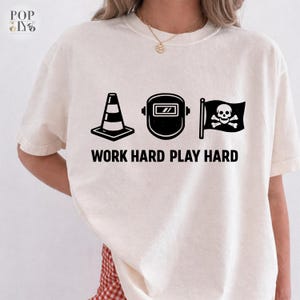 May include: Cream-coloured t-shirt with black graphics of a traffic cone, welding mask, and a skull and crossbones flag. The text "WORK HARD PLAY HARD" is printed below. The shirt has a relaxed fit.