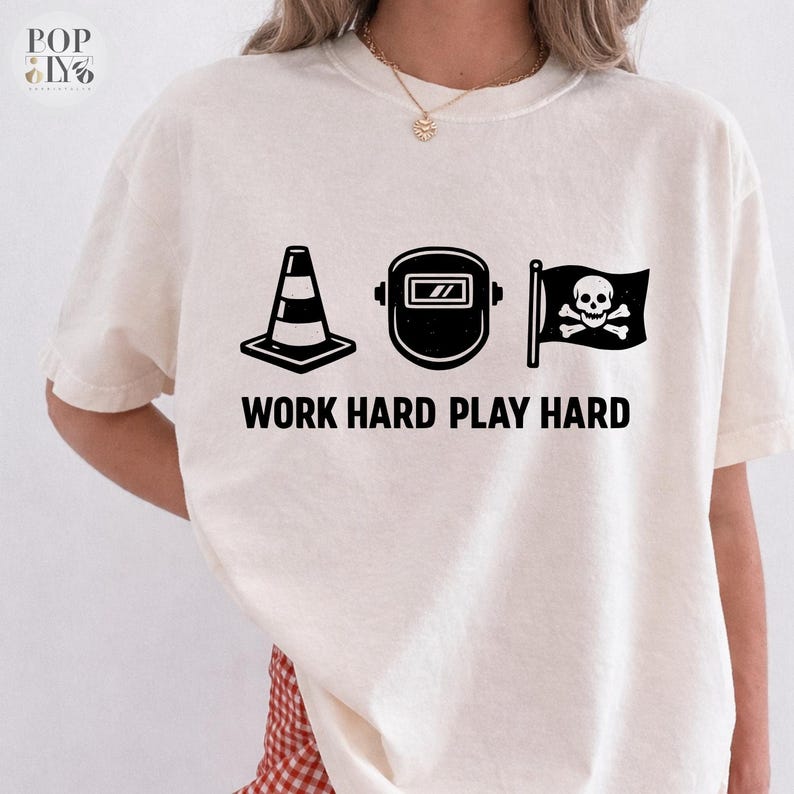 May include: Cream-coloured t-shirt with black graphics of a traffic cone, welding mask, and a skull and crossbones flag. The text "WORK HARD PLAY HARD" is printed below. The shirt has a relaxed fit.