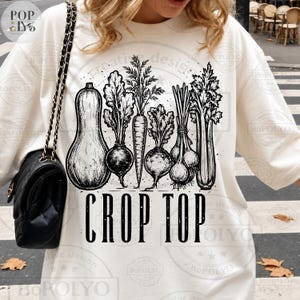 May include: A cream-coloured crop top with a black and white vegetable illustration, including a squash, beetroot, carrots, and onions. The words "CROP TOP" are printed in bold, black letters. A comfortable and stylish top.