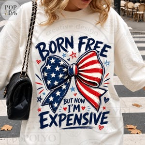 May include: White crew neck sweatshirt with a patriotic bow graphic. The bow features a design of the American flag with the text "BORN FREE BUT NOW I'M EXPENSIVE". The design includes stars, hearts, and a patriotic color scheme.