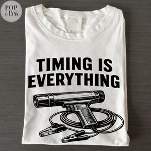 Puede incluir: Camiseta blanca con el texto "TIMING IS EVERYTHING" en negrita negra. Debajo, una ilustración en blanco y negro de una lámpara estroboscópica con cables y pinzas.