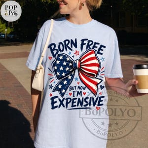 May include: Light blue t-shirt with a patriotic bow design in red, white, and blue. The shirt features the text "BORN FREE BUT NOW I'M EXPENSIVE" in a playful font. A coffee cup is held in the hand.