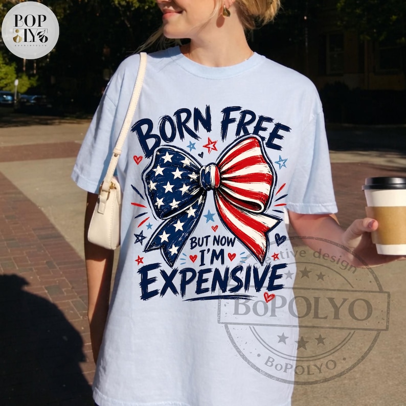 May include: Light blue t-shirt with a patriotic bow design in red, white, and blue. The shirt features the text "BORN FREE BUT NOW I'M EXPENSIVE" in a playful font. A coffee cup is held in the hand.