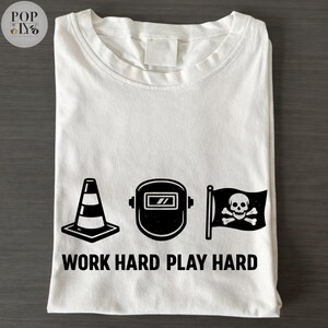 May include: White t-shirt with a black graphic design featuring a traffic cone, welding mask, and a skull and crossbones flag. The text "WORK HARD PLAY HARD" is printed below the graphics.