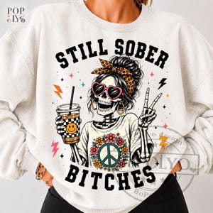 May include: A light gray crewneck sweatshirt featuring a graphic of a skeleton wearing heart-shaped sunglasses, a leopard print headband, and a floral peace sign t-shirt. The skeleton holds a coffee cup and makes a peace sign with text STILL SOBER BITCHES.