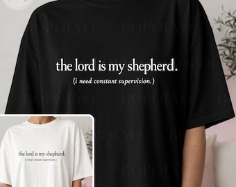 May include: Black t-shirt with the text "the lord is my shepherd." in a white, simple font. Below the text is the phrase "(i need constant supervision.)" in a smaller font. The image also shows a white t-shirt with the same text.